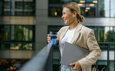Sales manager standing with laptop and coffee on office terrace during break time and looks away